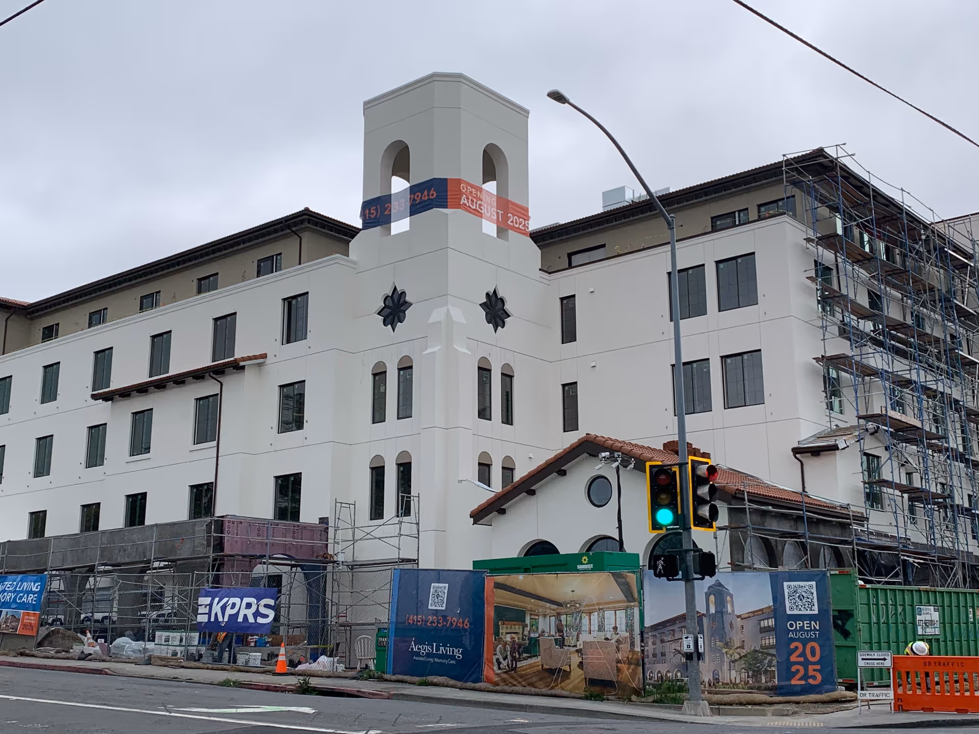 A large multi-story building under construction with scaffolding on one side and a tower-like structure in the center. The building has a white exterior with multiple windows and a tiled roof section. Construction fencing surrounds the site with banners displaying the name Aegis Living and an opening date of August 2025. A traffic light is visible in the foreground.