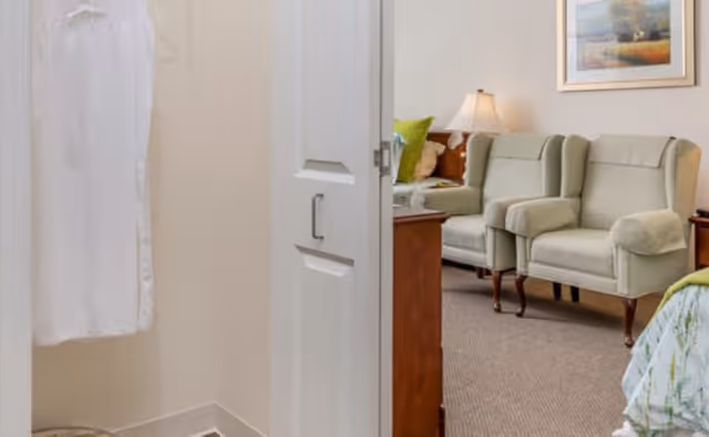 View from a small closet or dressing area with a white robe hanging on a hook, looking into a cozy living room area with two light green upholstered armchairs, a wooden side table with a lamp, and a framed landscape painting on the wall.