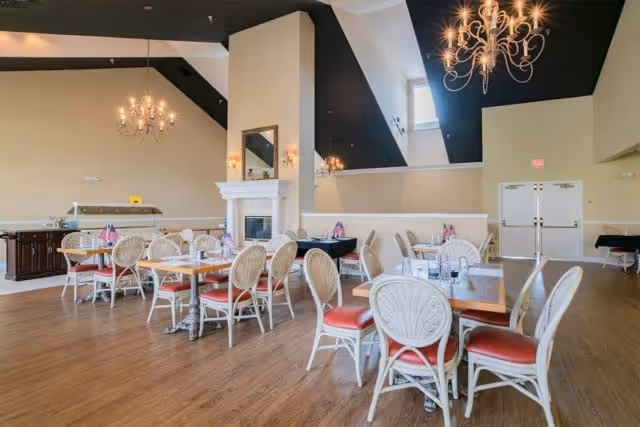 A spacious dining room with multiple wooden tables and white wicker chairs with red cushions. The room features a high ceiling with black accents and elegant chandeliers. There is a fireplace with a mirror above it, and a buffet area with a glass sneeze guard on the left side. The floor is wooden, and there are double doors at the far end of the room.