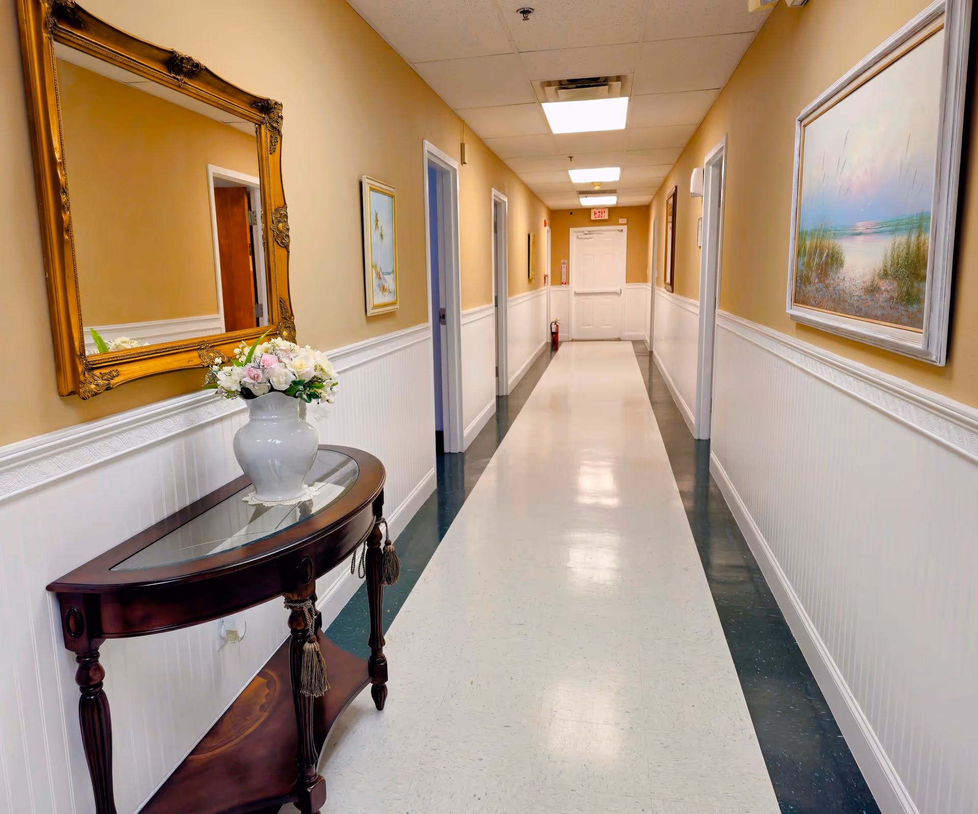 A well-lit hallway in an assisted living facility with beige walls and white wainscoting. The floor is white with dark green borders. On the left side, there is a wooden console table with a glass top holding a white vase filled with flowers, and above it hangs a large ornate gold-framed mirror. On the right wall, there is a framed painting of a beach scene. Several doorways line the hallway, and a white door is visible at the end.