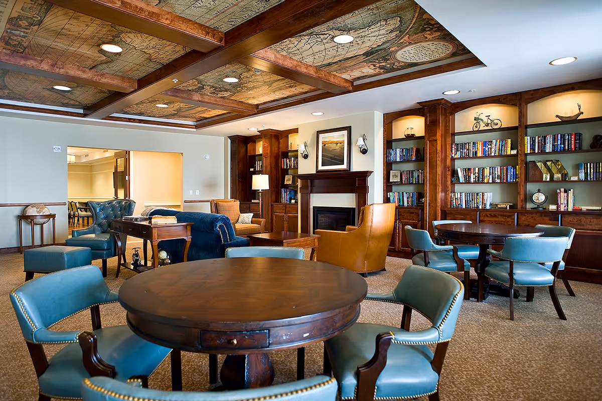 A cozy senior living facility common area with round wooden tables surrounded by teal upholstered chairs. The room features a fireplace with a framed picture above it, built-in wooden bookshelves filled with books and decorative items, and comfortable armchairs in blue and tan. The ceiling has wooden beams with a vintage map design. The space is well-lit with recessed lighting and a floor lamp.