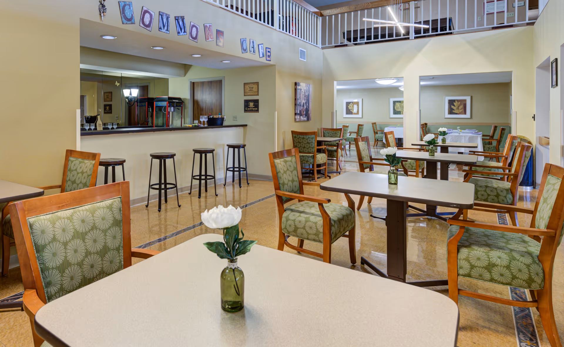 A bright and spacious dining area in a senior living community with multiple tables and chairs arranged neatly. Each table has a small vase with a white flower. There is a counter with bar stools and a popcorn machine in the background. The walls are painted light yellow and decorated with framed artwork and a sign that reads 'COMMON CAFE'.