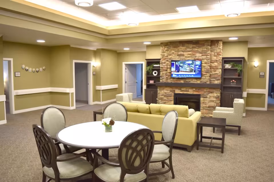 A spacious living room area with a round table and four chairs in the foreground. Behind the table is a yellow sofa and two armchairs facing a stone fireplace with a mounted flat-screen TV. Built-in shelves flank the fireplace, and there are several doorways leading to other rooms. The walls are painted light green and the ceiling has recessed lighting.