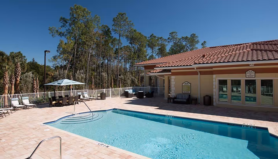 Outdoor swimming pool area with lounge chairs, tables with umbrellas, and a building with a tiled roof in the background surrounded by trees under a clear blue sky.
