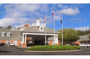 Front entrance of a brick senior living community with a covered porte-cochere, three flagpoles on a circular driveway, and surrounding landscaping.
