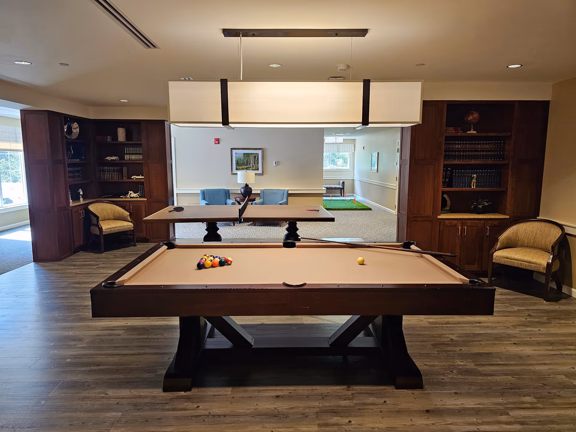 A recreational room featuring a pool table with billiard balls arranged for a game. Behind the pool table is a table with two blue chairs and a lamp, and further back is a small indoor putting green. The room has wooden bookshelves with decorative items and chairs on either side, with wood flooring and soft lighting.