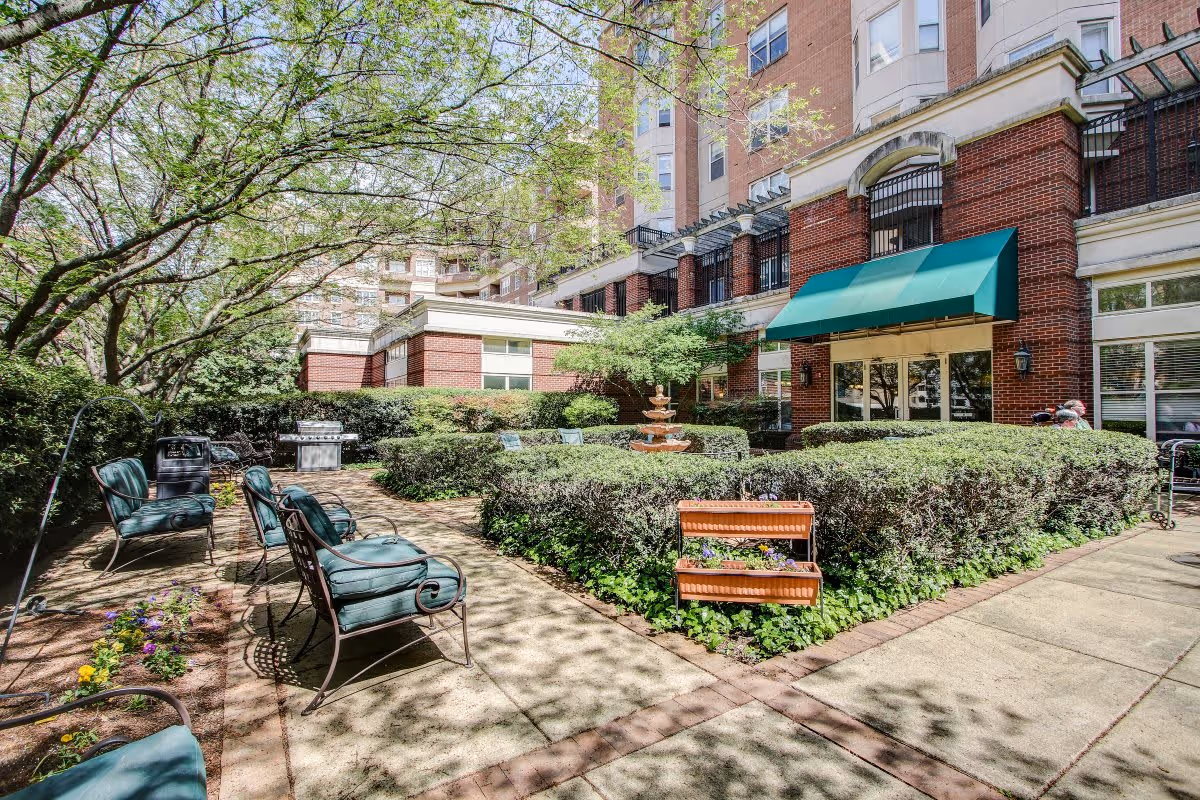 Outdoor courtyard area of a senior living facility with green cushioned metal chairs, a small fountain, neatly trimmed hedges, flower beds, and a brick building with a green awning in the background.