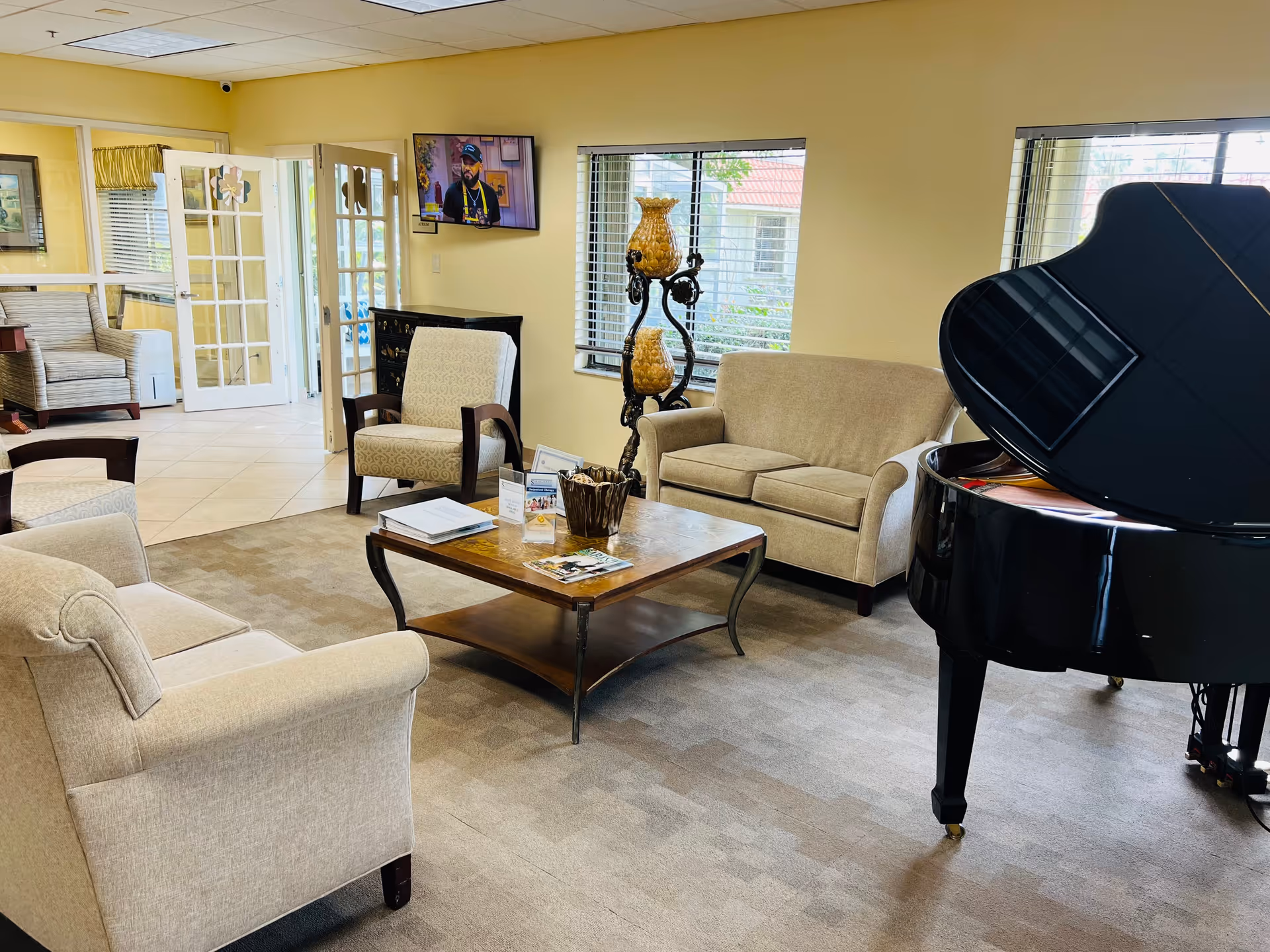 Well-lit senior living lounge with sofas, armchairs, a coffee table and a grand piano near windows.