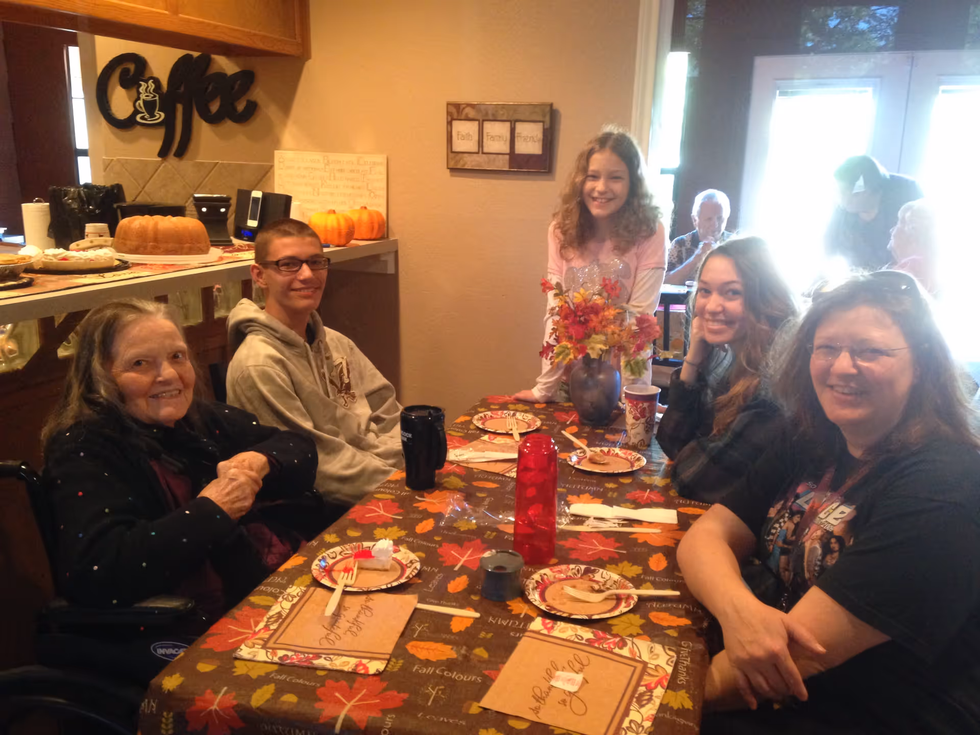 A group of five people, including elderly and younger individuals, sitting and standing around a dining table decorated with a fall-themed tablecloth and a vase of autumn flowers. Plates with food and drinks are on the table. The background shows a kitchen area with a coffee sign and some baked goods.