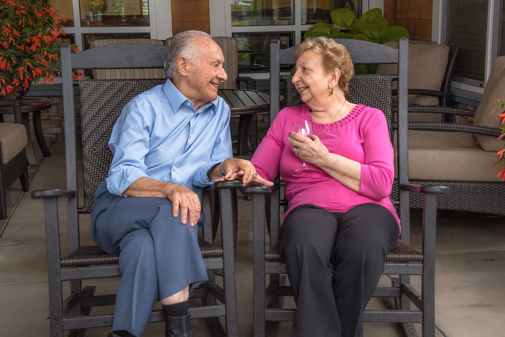 An elderly man and woman sitting on rocking chairs on a patio, holding hands and smiling at each other. The woman is holding a glass of wine. There are plants and outdoor furniture around them.