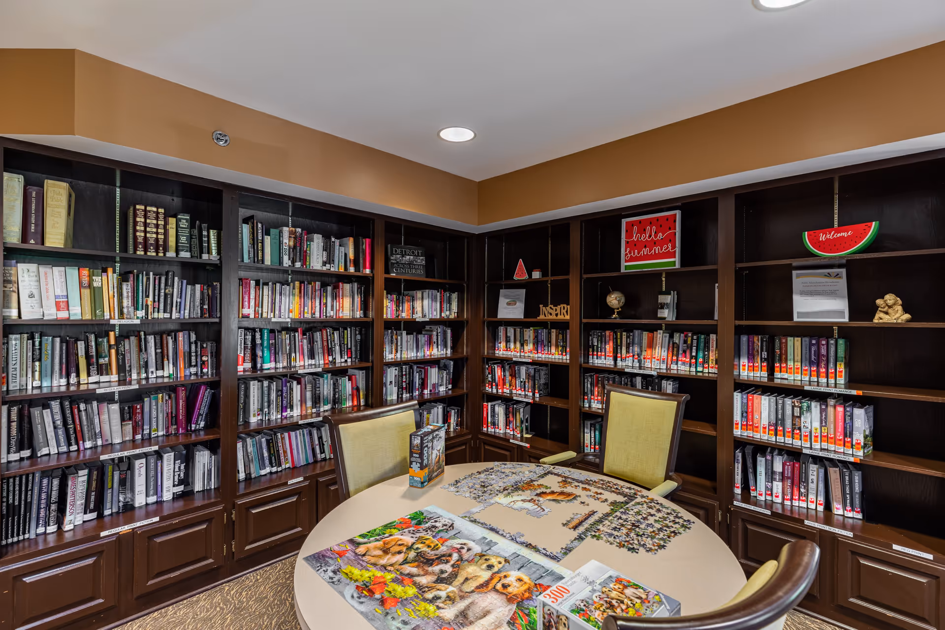 A cozy library room with dark wooden bookshelves filled with books lining the walls. In the center, there is a round table with a partially completed dog-themed jigsaw puzzle and two puzzle boxes. Two cushioned chairs are positioned around the table. The room has warm lighting and a welcoming atmosphere with small decorative signs on the shelves.