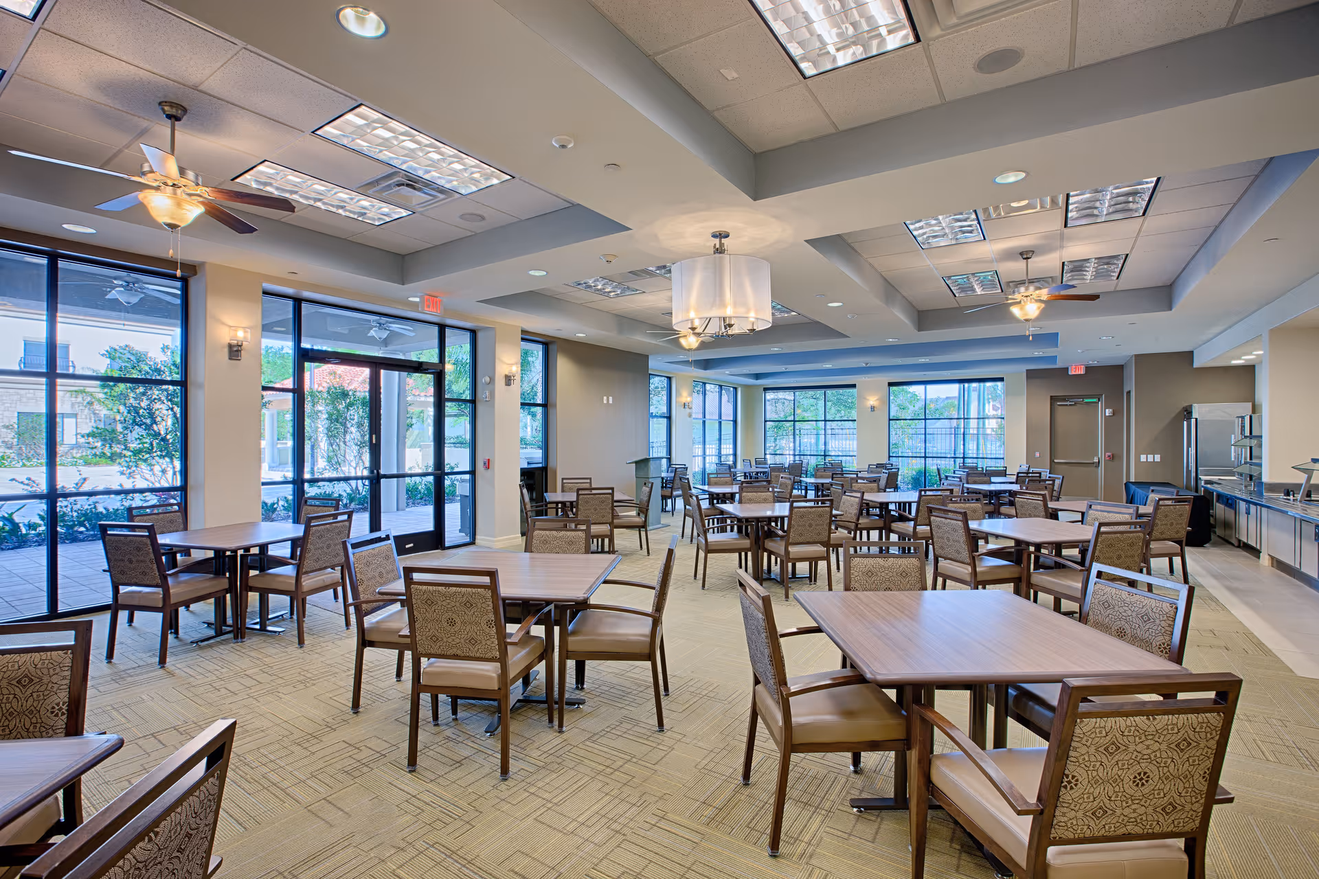 A spacious dining room in a senior living facility with multiple wooden tables and cushioned chairs arranged neatly. Large windows allow natural light to fill the room, and ceiling fans and modern light fixtures hang from the ceiling. The room has a neutral color palette with beige walls and carpeted floors.