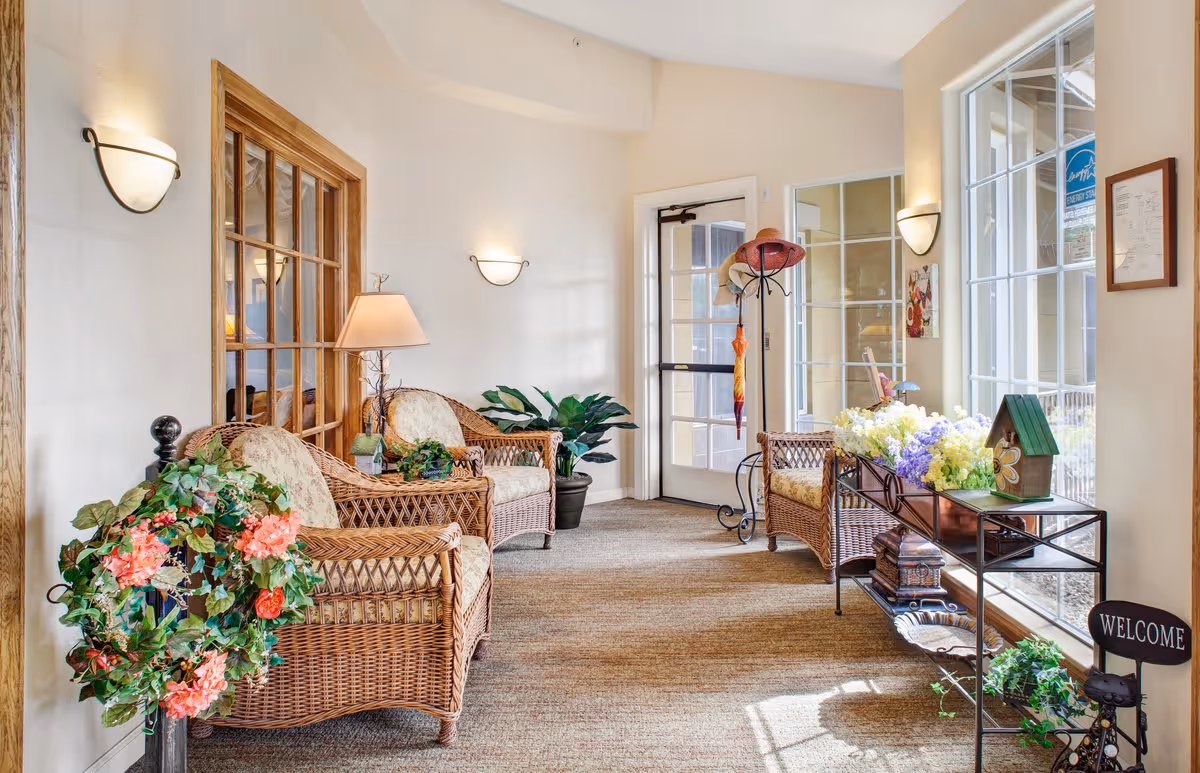 Sunlit seating area with wicker chairs, plants, floral arrangements and a welcome sign near large windows and a glass door.