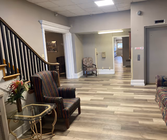 A senior living facility seating area with upholstered armchairs, a small glass side table with flowers, a staircase, and a hallway leading past an elevator.