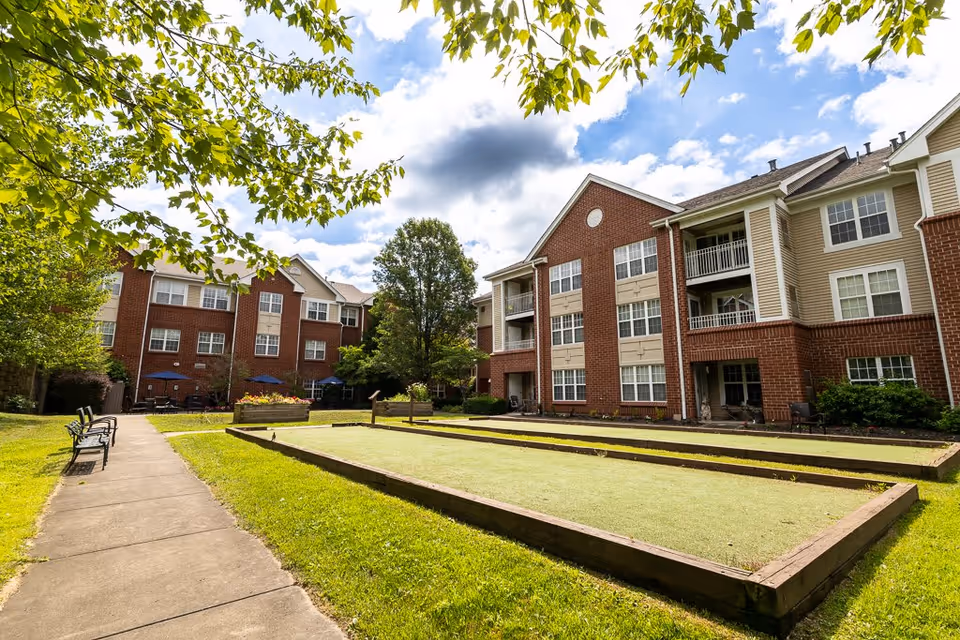 Outdoor view of Elison Independent & Assisted Living of Maplewood showing a well-maintained lawn with bocce ball courts, benches along a paved walkway, and multi-story residential buildings with brick and beige siding under a partly cloudy sky.