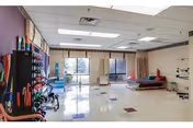 A spacious rehabilitation therapy room with exercise equipment including therapy balls, parallel bars, and mats. The room has large windows allowing natural light, white walls, and a tiled floor with some colored tiles.