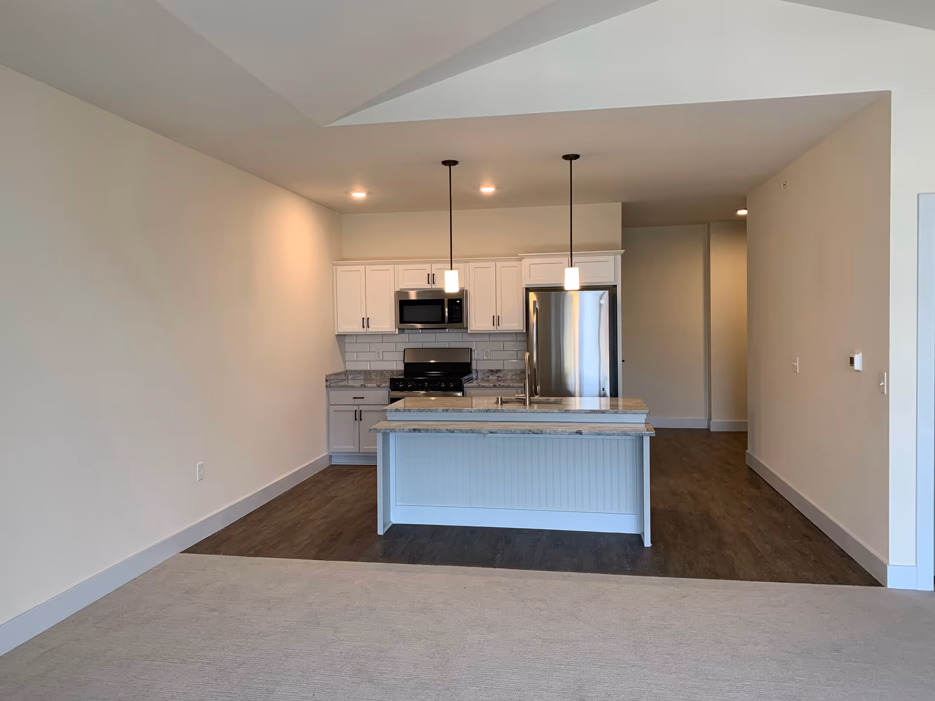 Modern kitchen area with white cabinets, stainless steel refrigerator, microwave, and stove. There is a kitchen island with a sink and two pendant lights hanging above it. The floor is a combination of wood and carpet, and the walls are painted light beige.