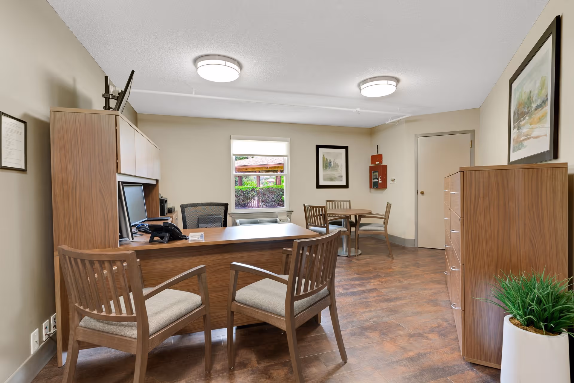 An office room with a wooden desk and two cushioned wooden chairs in front of it. Behind the desk is an office chair, a computer monitor, and a telephone. There is a window with a view of greenery outside, a small round table with three chairs, a wooden cabinet with drawers, a potted plant, and framed artwork on the walls. The room has beige walls and wood-patterned flooring with two ceiling lights.