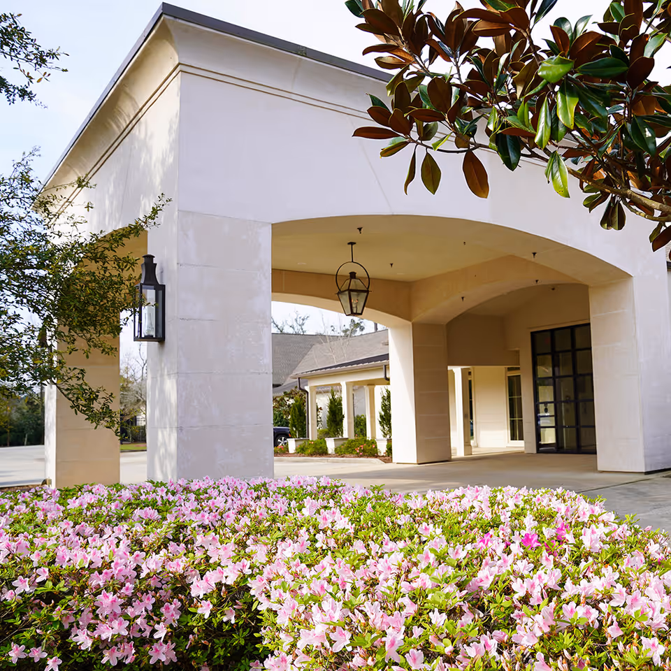 Entrance of The Peristyle at Beau West facility featuring a covered driveway with large beige columns, hanging lantern-style lights, and a bed of blooming pink flowers in the foreground. Trees with green leaves frame the scene.