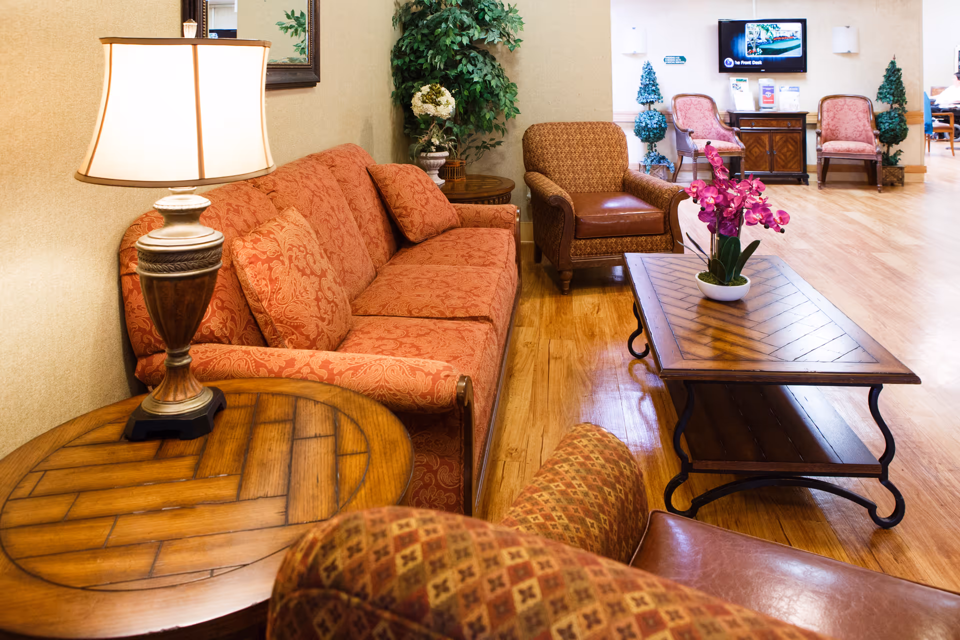 A cozy living room area with a patterned red sofa, matching armchairs, a wooden coffee table with a purple orchid centerpiece, and a side table with a lamp. The room has wooden flooring and light-colored walls, with some greenery and additional seating visible in the background.