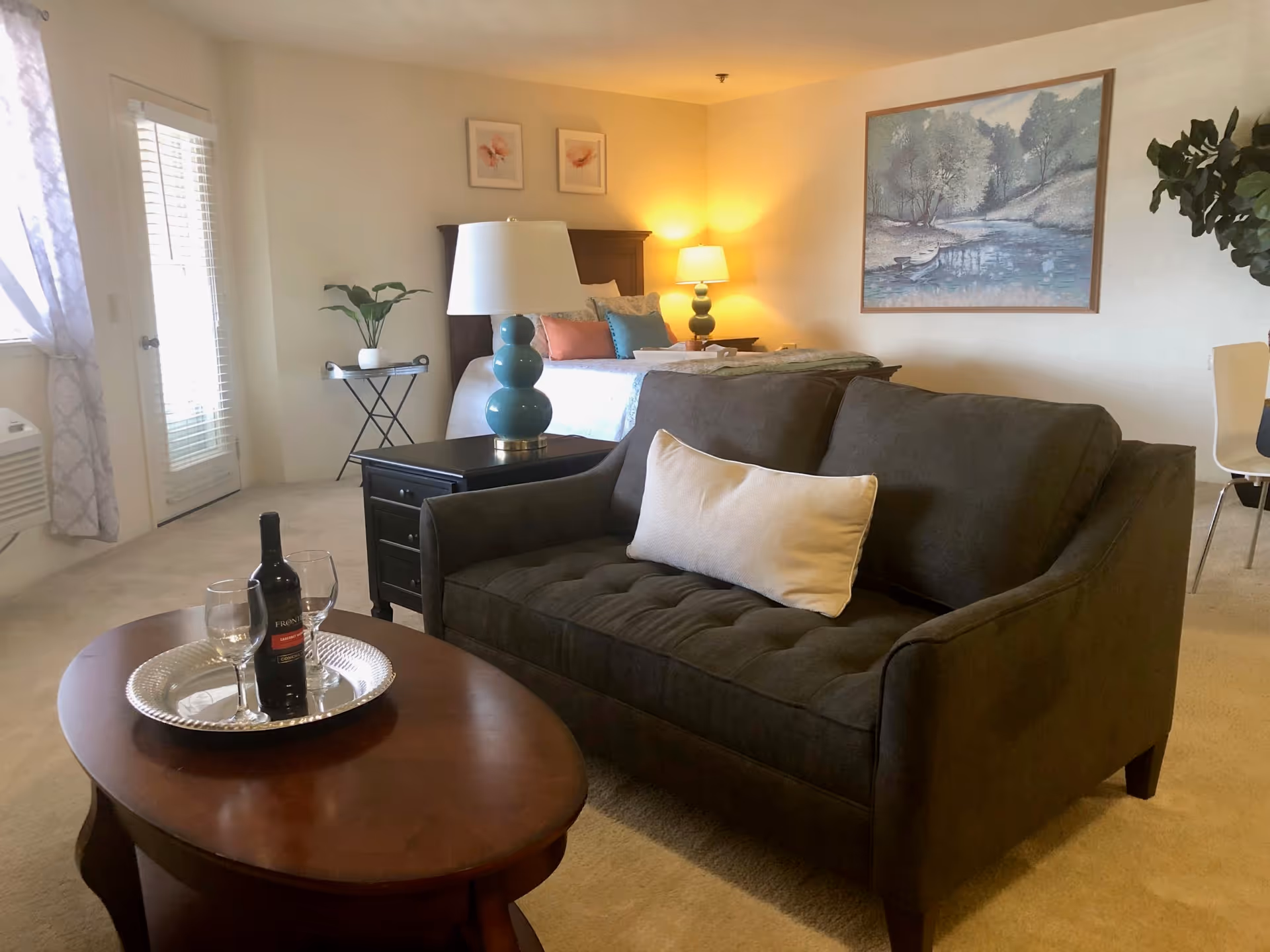 Cozy studio living area with a dark loveseat, coffee table holding a bottle of wine and glasses, and a bed with lamps in the background.