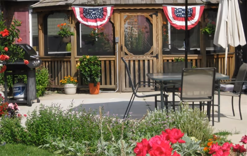 Outdoor patio area with a table and chairs, a barbecue grill, and various potted plants and flowers. The background shows a wooden screened porch decorated with red, white, and blue bunting.