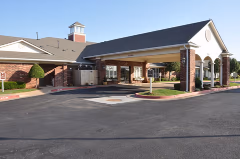 Front entrance of a brick senior living facility with a covered porte-cochere, circular driveway, and landscaped shrubs.
