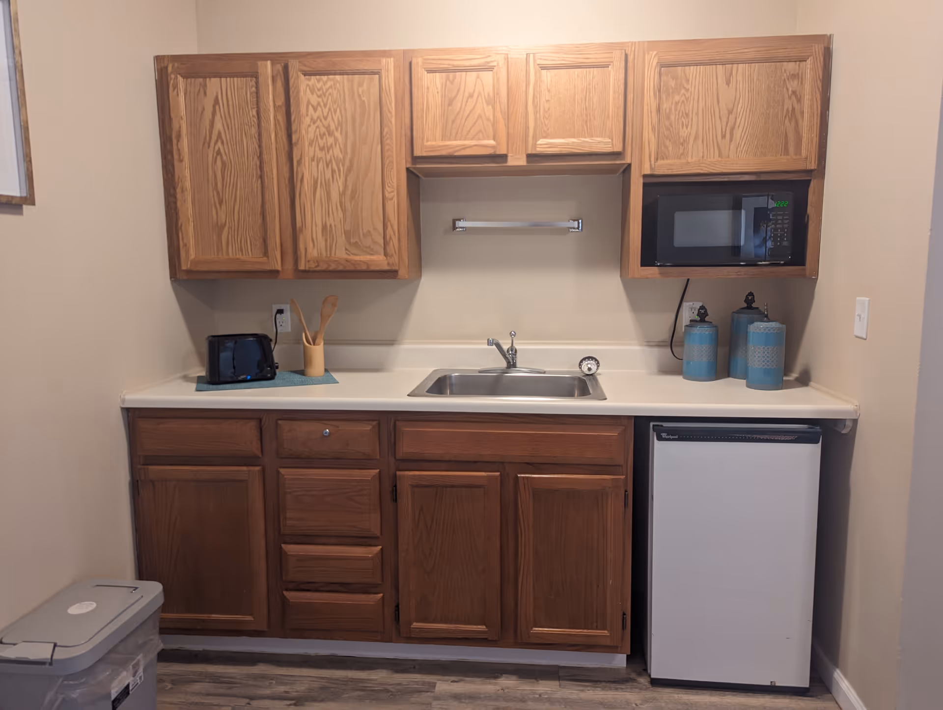 A small kitchen area with wooden cabinets, a countertop with a sink, a black toaster, wooden utensils in a holder, a microwave, two blue decorative canisters, and a compact refrigerator. The floor has a wood-like finish and there is a trash bin on the left side.