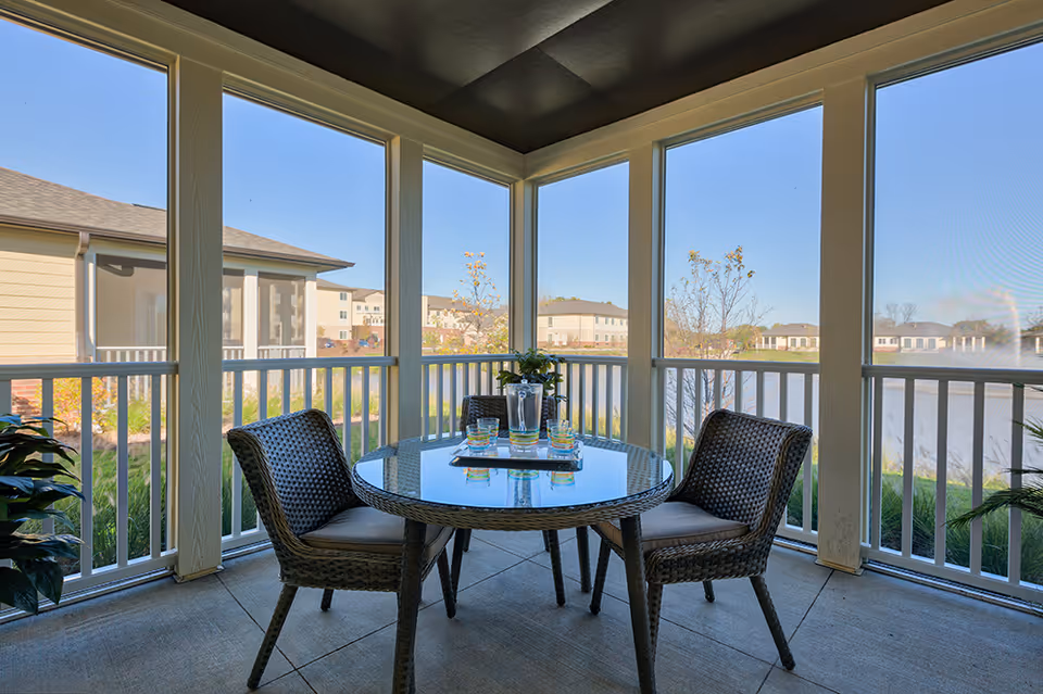 A screened-in outdoor patio area with a round glass-top table and four wicker chairs. The table has a tray with a pitcher and four glasses. The patio overlooks a landscaped area with grass, trees, and a pond, with residential buildings visible in the background under a clear blue sky.