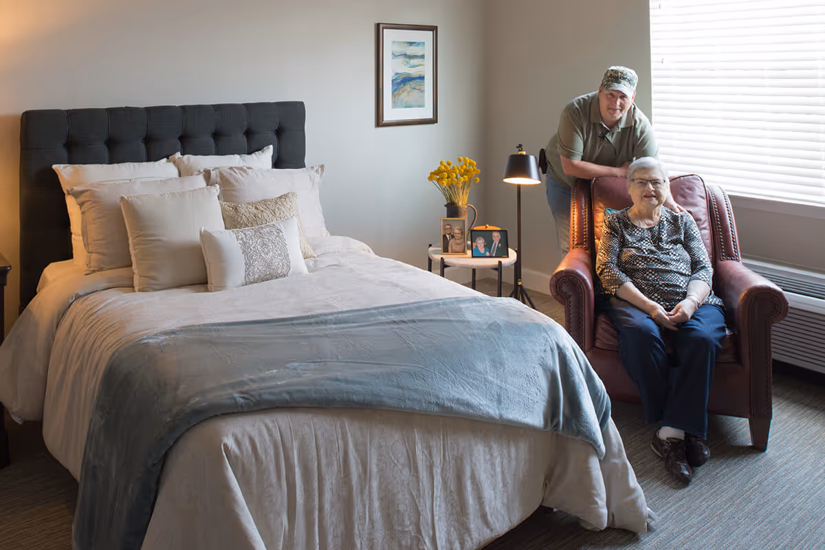A cozy bedroom with a neatly made bed featuring multiple pillows and a light blue throw blanket. An elderly woman is sitting in a brown armchair near a window with blinds, and a man wearing a cap is standing behind her, smiling. A small round table next to the chair holds a vase with yellow flowers and framed photographs, with a floor lamp providing additional light.