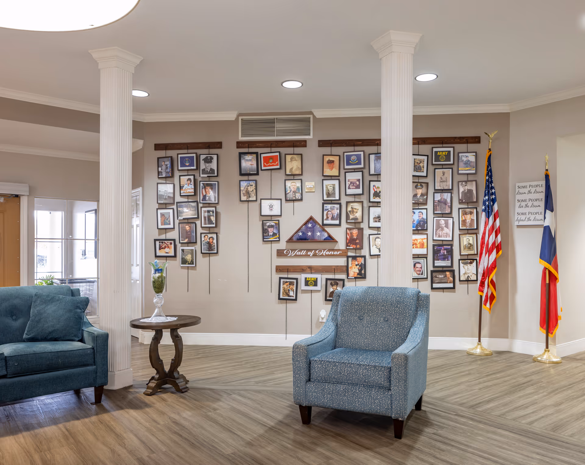 A seating area in a senior living facility with two armchairs and a small round wooden table between two white columns. Behind the seating area is a 'Wall of Honor' displaying numerous framed photographs and a folded American flag. To the right, there are two flags, one American and one Texas state flag, along with a sign that reads 'Some people dream the dream, some people live the dream, some people defend the dream.' The room has wood flooring and neutral-colored walls.