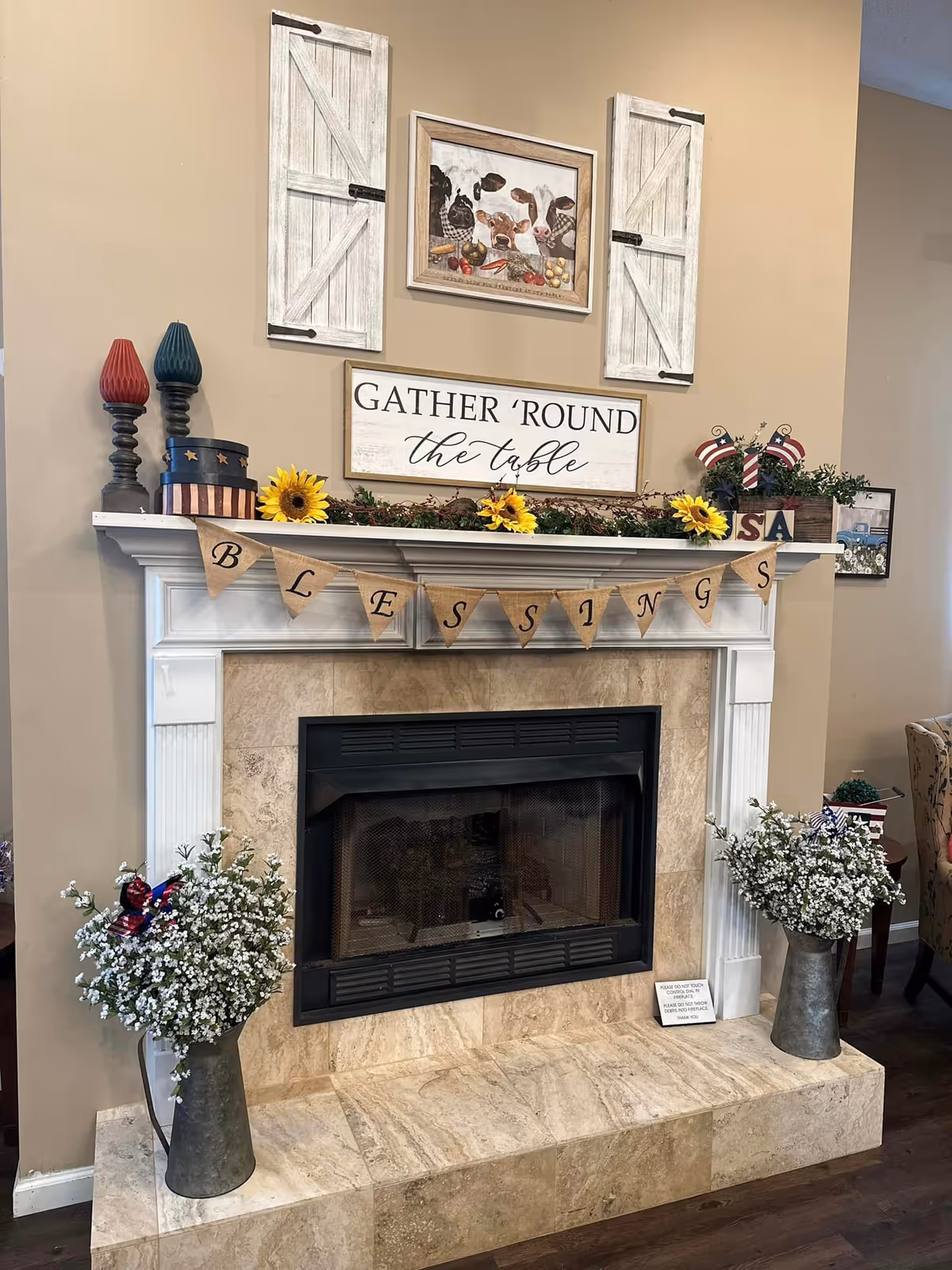 A cozy fireplace with a white mantel decorated with a garland of sunflowers and greenery. Above the mantel, there is a sign that reads 'GATHER 'ROUND the table' and a framed picture of cows. Two white rustic shutter-style wall decorations flank the picture. A burlap banner spelling 'BLESSINGS' hangs across the mantel. On either side of the fireplace, there are metal vases filled with white flowers and small patriotic decorations. The fireplace surround is made of beige stone tiles.