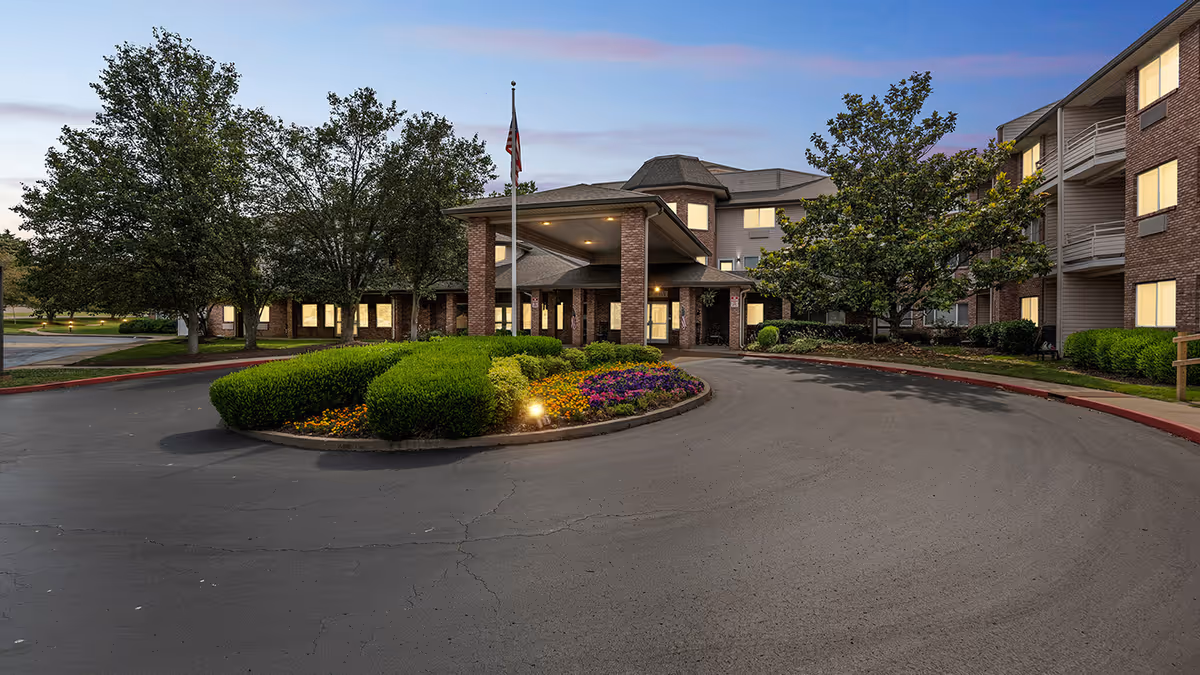 Entrance of a multi-story senior living building with a circular driveway, landscaped island, flagpole and illuminated windows at dusk.