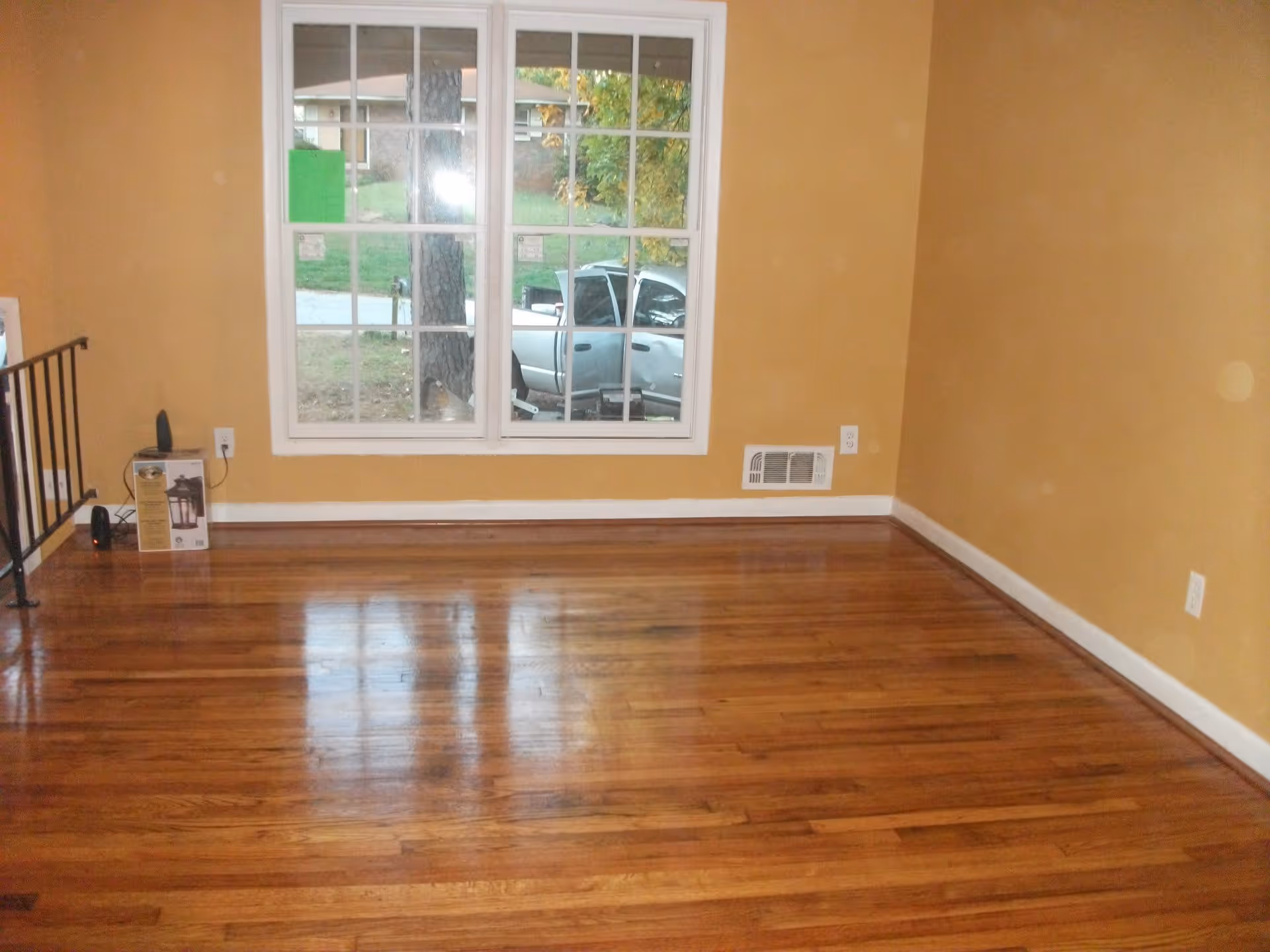 Empty room with polished wooden floor, mustard yellow walls, a large window showing a parked silver truck outside, and a small box and speaker near the left wall.