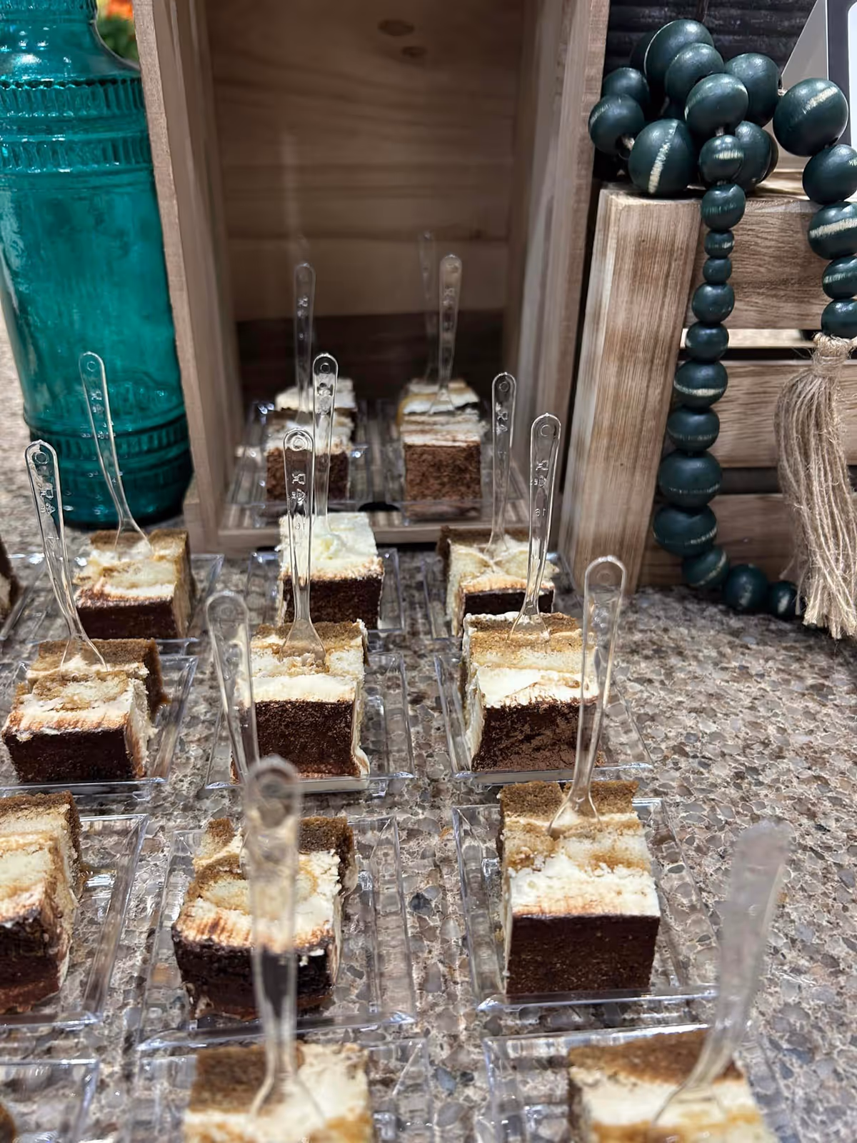 Small square dessert bites in clear plastic dishes with tiny spoons arranged on a countertop, with wooden decor and a green beaded garland in the background.