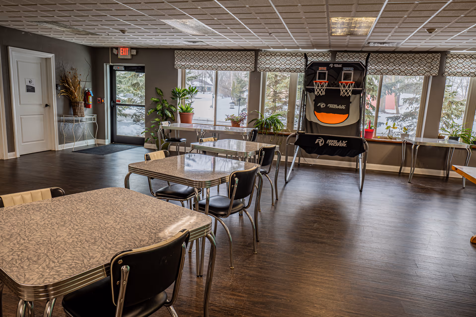 A bright communal dining/activity room with multiple retro-style tables and chairs, potted plants by large windows, and a standing basketball arcade game.