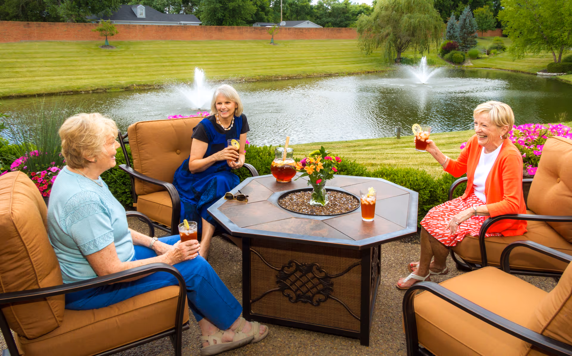 Three elderly women sitting outdoors on cushioned patio chairs around a hexagonal table with drinks, flowers, and a pitcher. They are smiling and enjoying a sunny day by a pond with two water fountains, surrounded by green grass, trees, and colorful flowers.