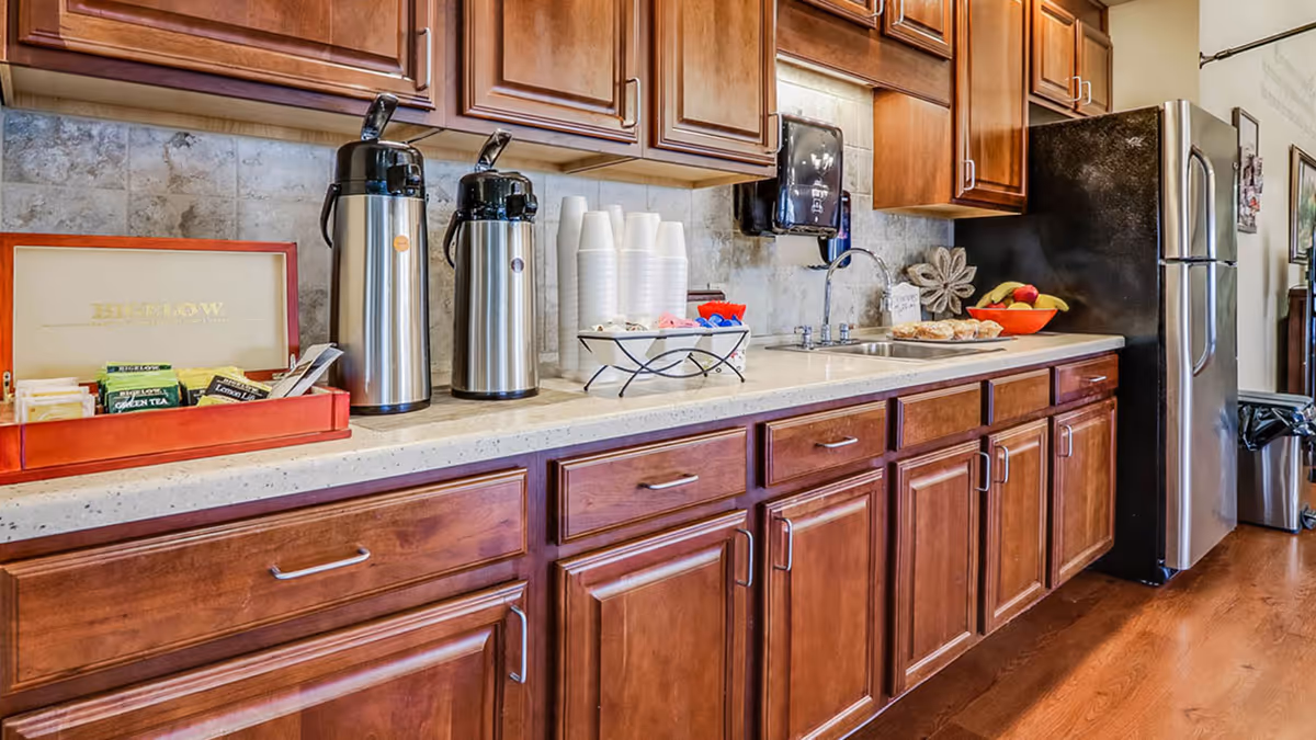 A kitchen area with wooden cabinets and drawers, a countertop with two stainless steel airpots, a red box containing various tea bags, a stack of white disposable cups, a small tray with creamers and sweeteners, a sink with a faucet, a bowl of fruit, and a stainless steel refrigerator.