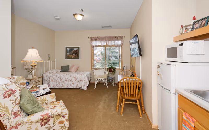 Studio apartment room with a bed, floral armchair, small kitchen area, dining table, and wall-mounted TV.