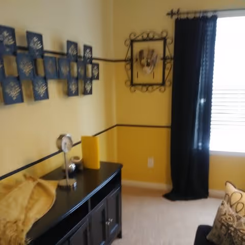 A cozy corner of a room with yellow walls and black trim, featuring a black cabinet with decorative items on top, a yellow throw blanket, a wall-mounted artwork, and a window with black curtains letting in natural light.