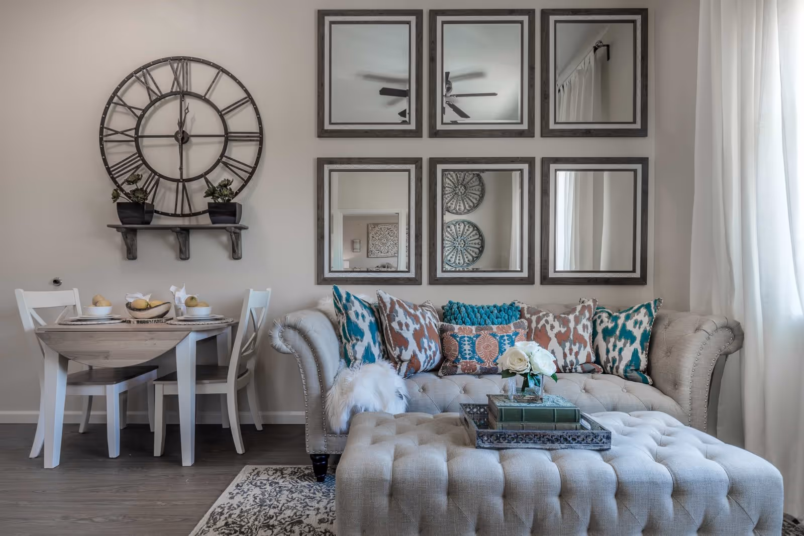 Stylish living room with a tufted sofa and ottoman, colorful throw pillows, six framed mirrors, a large wall clock, and a small dining table.