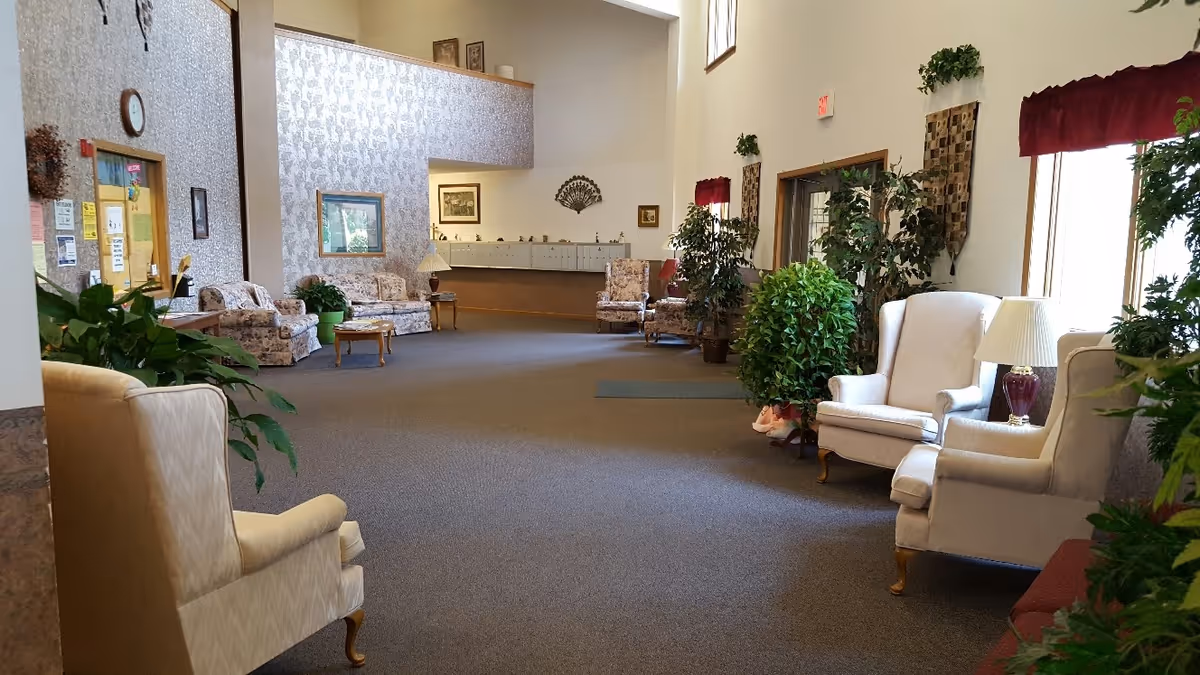 A spacious and well-lit common area in a senior living facility featuring several upholstered armchairs and sofas arranged along the walls. The room has carpeted flooring, potted plants, framed artwork on the walls, and large windows with red valances allowing natural light to enter. A reception or mail area is visible in the background.