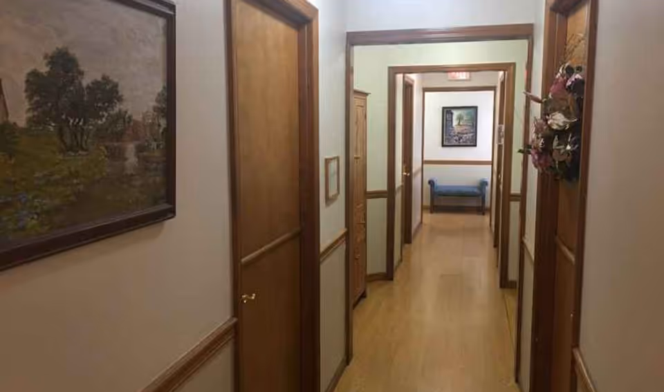 A wood-trimmed corridor in a care facility with doors on both sides, framed paintings, and a bench at the end.