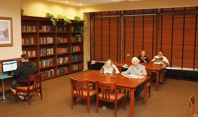 A quiet reading room in a retirement community with wooden tables and chairs. Four elderly people are seated at two tables, reading books and newspapers. A man is using a computer at a small desk on the left side of the room. There is a large bookshelf filled with books against the wall, and the windows have closed wooden blinds.