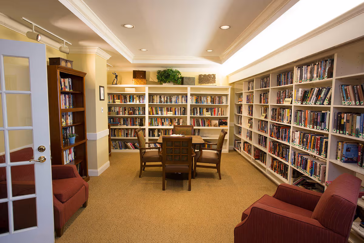 Well-lit interior library with bookshelves lining the walls, a central table and chairs, and upholstered armchairs near the entrance.