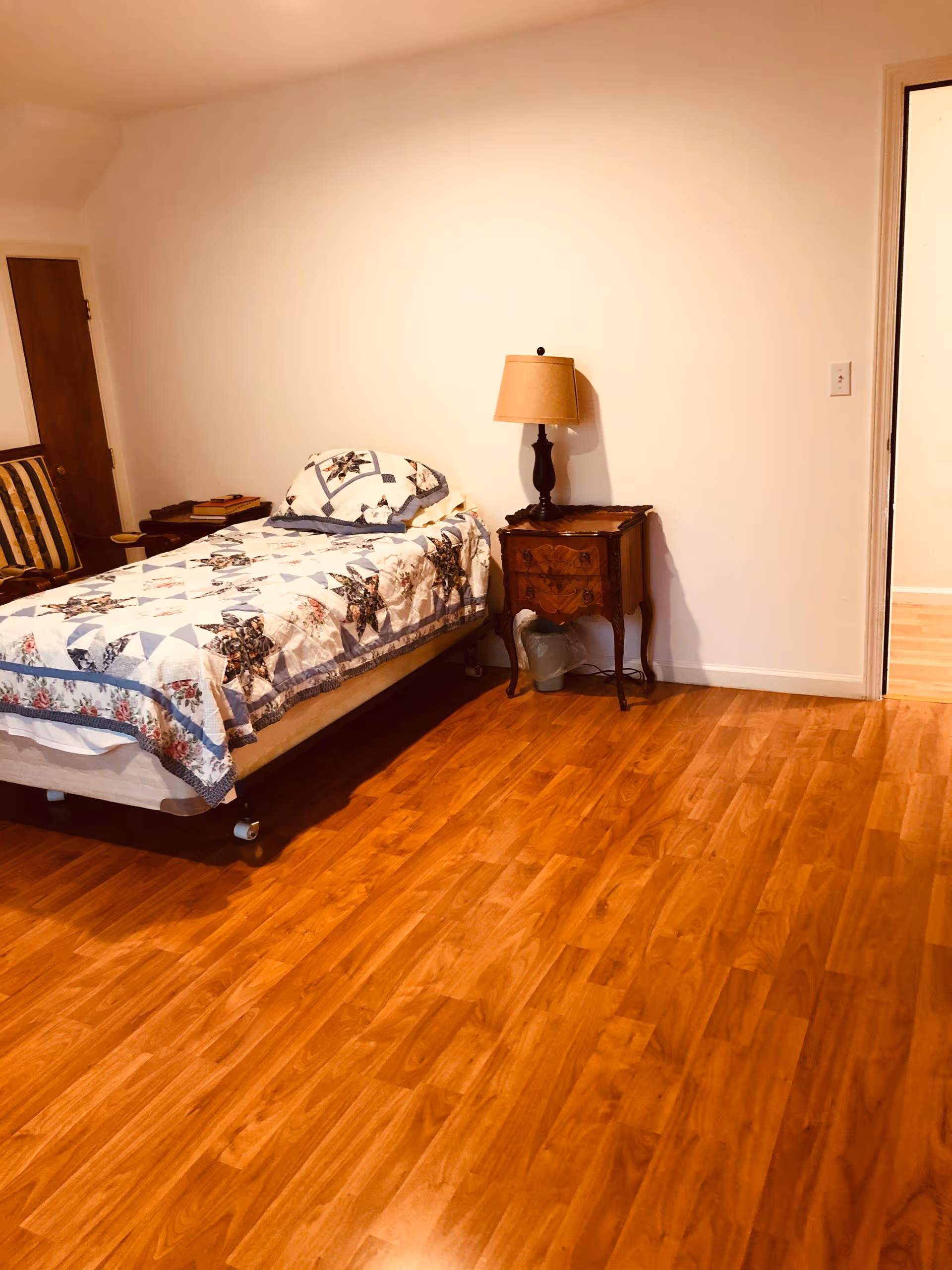 A simple bedroom with a single bed covered in a patterned quilt, a wooden nightstand with a lamp, a wooden chair with striped upholstery, and hardwood flooring. The walls are plain white, and there is an open doorway leading to another room.