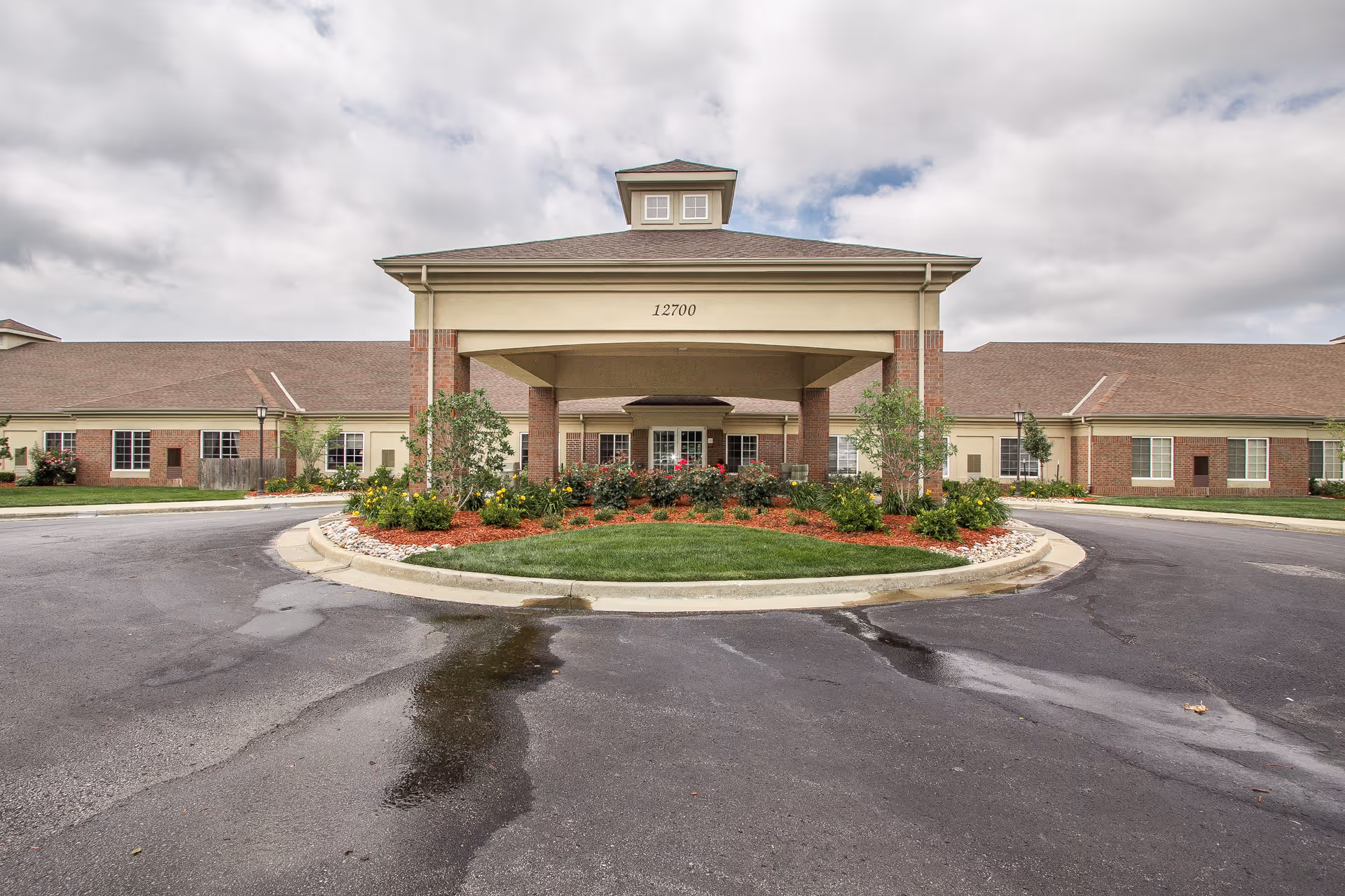 Front exterior view of a single-story senior living facility building with a covered entrance, landscaped garden with flowers and shrubs, and a circular driveway under a cloudy sky.