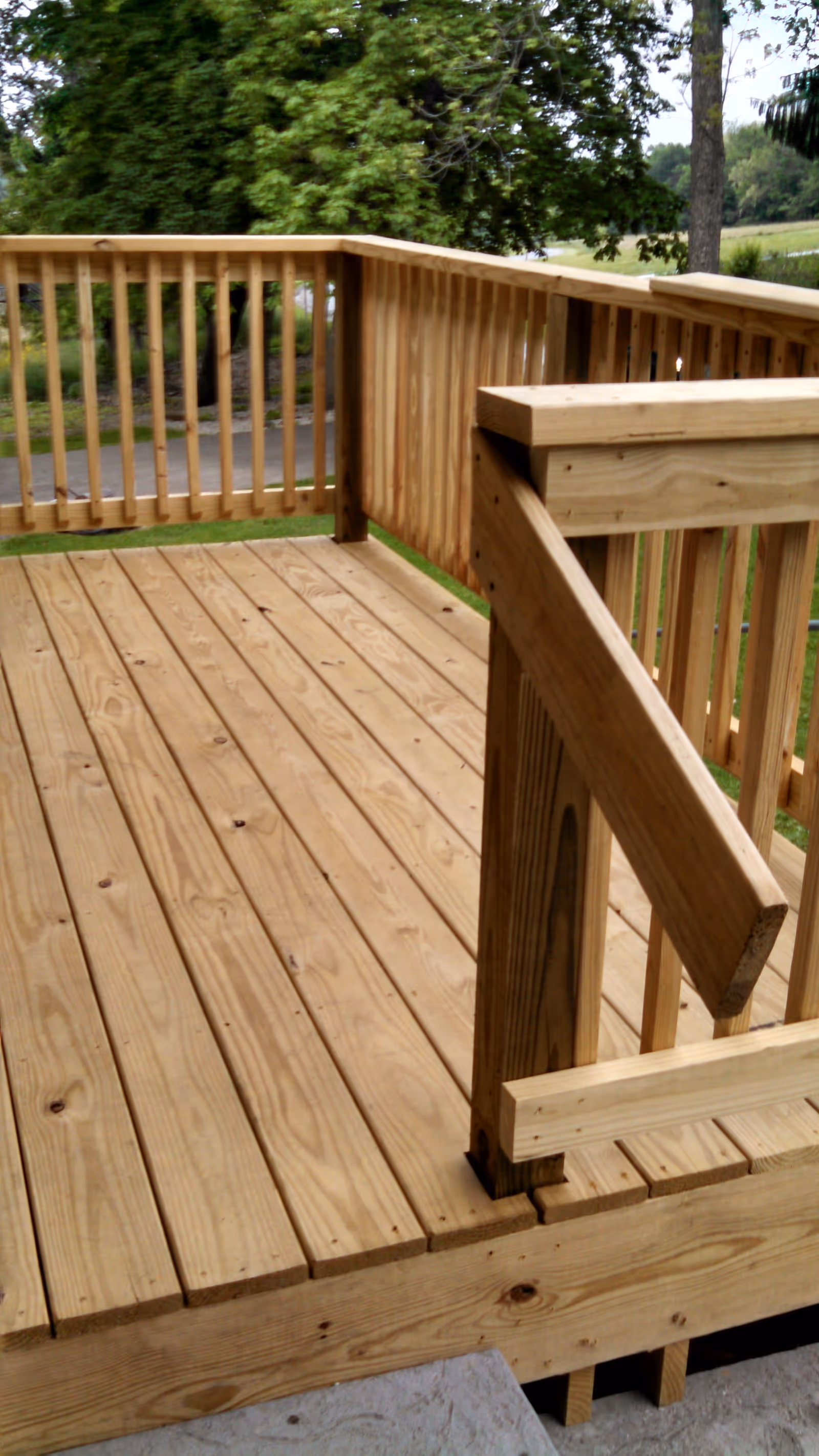 A wooden outdoor deck with railings, surrounded by green trees and grass in the background.