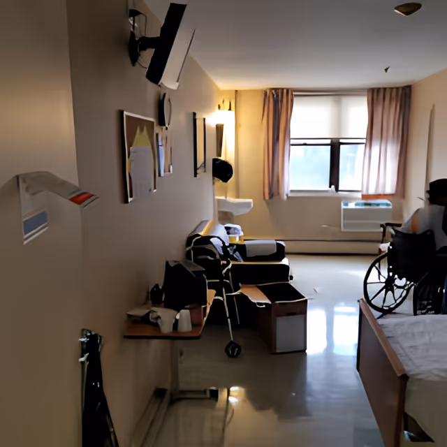 Interior view of a senior living facility room with a wheelchair, armchairs, a walker, a bed, and a window with curtains letting in natural light. The room has a beige wall with some pictures and a mounted television.