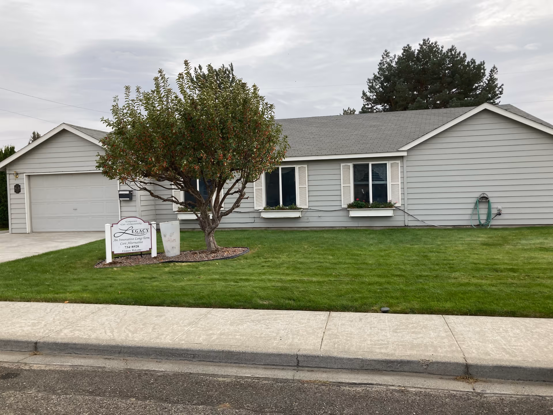 Single-story gray house with white trim and a two-car garage. There is a tree with green leaves in the front yard, which is well-maintained with green grass. A sign near the tree reads 'Legacy An Innovative Long-Term Care Alternative'. The sky is overcast.